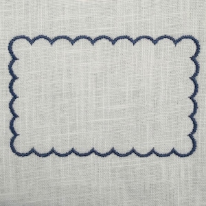 Op de afbeelding: Witte linnen stof met een marineblauwe, geschulpte geborduurde rand. De geborduurde rechthoek is gecentreerd op de stof, wat een decoratief element creëert. De stof heeft een subtiele textuur.