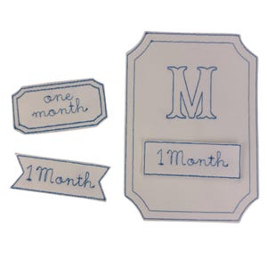 May include: A set of four white fabric patches with blue stitched details. The patches include the text "one month" and "1 Month". One patch features a large blue letter "M".