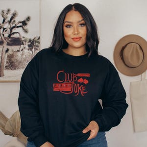 May include: Black sweatshirt with the red text "Club Juke" and a graphic of a piano keyboard, a harmonica, and playing card suits. The sweatshirt is a crew neck style.