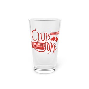 Pode incluir: Um copo de cerveja transparente com um gráfico vermelho que diz "Club Juke". O design inclui um teclado de piano, uma pauta musical e símbolos de cartas de baralho. O copo está vazio e está sobre uma superfície branca.
