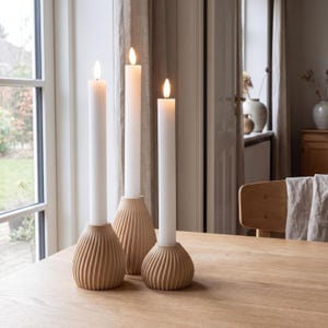 Peut inclure: Trois bougies blanches avec des flammes vacillantes sont placées dans des bougeoirs beiges et côtelés. Les bougeoirs sont disposés sur une table en bois clair, créant une ambiance chaleureuse. La scène est éclairée par la lumière naturelle.