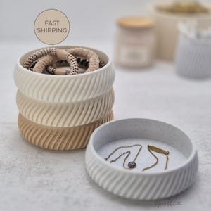 Pode incluir: Uma pilha de três recipientes redondos decorativos e texturizados em creme, bege e castanho. O recipiente superior contém elásticos de cabelo. Um recipiente semelhante separado contém dois colares. A imagem inclui o texto "FAST SHIPPING".