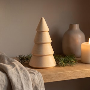 Puede incluir: Una decoración de árbol de Navidad de madera con un diseño en capas. El árbol es de color marrón claro y se encuentra sobre una estantería de madera con una ramita de hoja perenne. Un jarrón beige y una vela encendida están en el fondo.