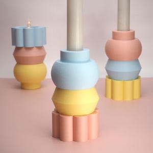 Puede incluir: Tres candelabros coloridos en tonos pastel de azul, rosa y amarillo. Los candelabros tienen un diseño geométrico con formas apiladas y están rematados con velas blancas. Los candelabros están sobre una superficie rosa.
