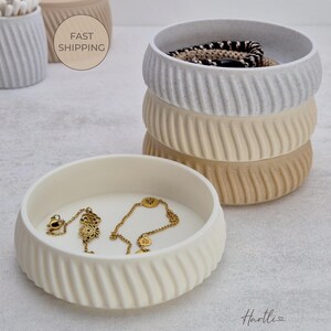 May include: A stack of three ceramic bowls in white, beige, and tan, with a ribbed exterior design. The bottom bowl holds gold jewelry. A fourth bowl is visible in the background, along with a container of cotton swabs.