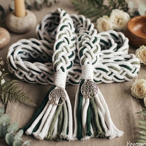 May include: A braided cord belt featuring a woven pattern of white, dark green, and olive green cotton strands. The ends are finished with white wrapped cord sections, hanging tassels, and circular silver-toned tree of life charms.