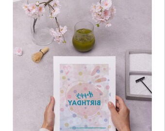May include: A birthday card with a colorful design and the words "Happy Birthday" in a teal font. The card is held by a person. The image also includes a glass of green liquid, cherry blossoms, and a matcha whisk.