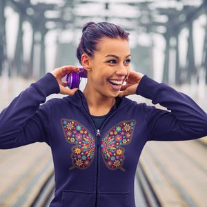 May include: A navy blue zip-up hoodie with a vibrant butterfly design on the front. The butterfly is composed of floral patterns in yellow, pink, blue, and green. The person is wearing purple headphones.