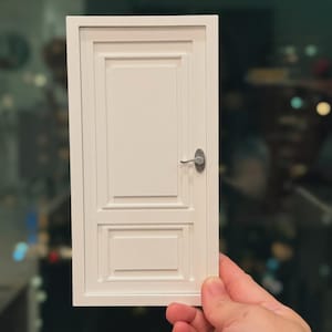 Może przedstawiać: Miniaturowe białe drzwi z srebrną klamką. Drzwi mają prostokątną ramę i dwa wpuszczane panele. Drzwi są trzymane w dłoni na rozmytym tle.