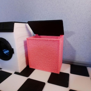 May include: A miniature laundry room scene featuring a white washing machine with a black door and a pink laundry hamper with a black lid. The scene is set on a black and white checkered floor.