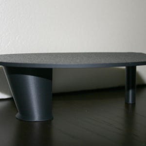 May include: A small, dark gray table with a textured surface. The table has a unique design, featuring a curved leg on one side and three cylindrical legs on the other. The table is approximately 24 inches long.