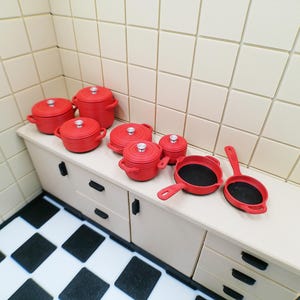 May include: A collection of miniature red cookware, including pots, pans, and lids, arranged on a light-colored cabinet. The set includes various sizes, with black interiors in the pans. The scene is set in a miniature kitchen with tiled walls and a checkered floor.