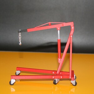May include: A red model engine hoist with a black boom and silver chain and hook. The hoist has a red hydraulic cylinder and is mounted on four small wheels. The background is a gradient of yellow and black.