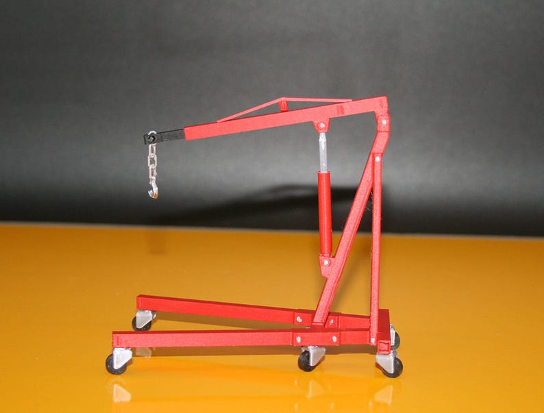 May include: A red model engine hoist with a black boom and silver chain and hook. The hoist has a red hydraulic cylinder and is mounted on four small wheels. The background is a gradient of yellow and black.