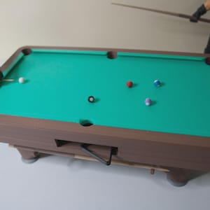 May include: A miniature pool table with a green felt surface and brown wooden frame. Several multi-colored billiard balls are scattered across the table. A cue stick is positioned to strike a white ball. A fun game for all ages.