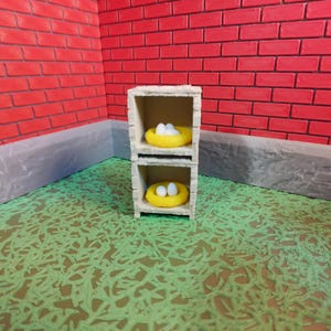 Peut inclure: Une petite étagère à deux niveaux avec deux nids jaunes, chacun contenant des œufs blancs. L'unité est placée contre un mur de briques rouges et un sol vert à motifs. La scène évoque un thème de ferme ou de printemps.