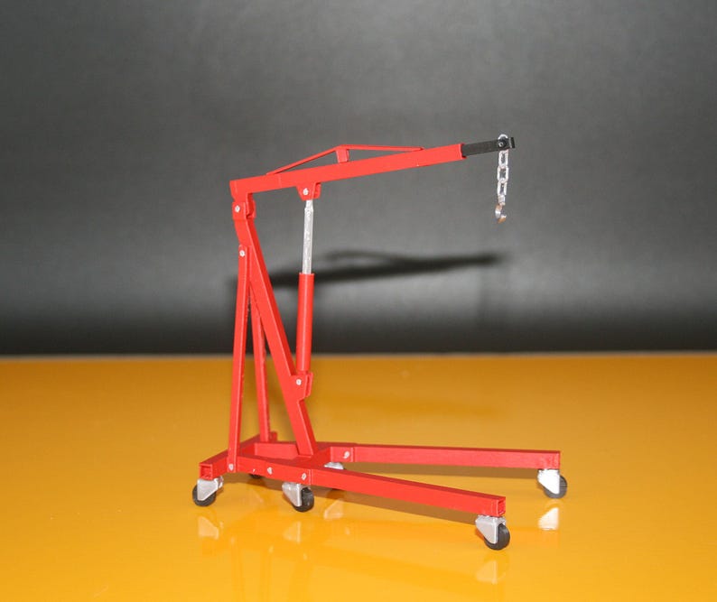 May include: A red model engine hoist with a silver chain and hook. The hoist has a wheeled base and a lifting arm. The background is a gradient of yellow and gray.