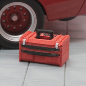 May include: A small, red toolbox with black accents and a black handle. The toolbox is sitting on a gray tiled surface, with a red car wheel in the background.