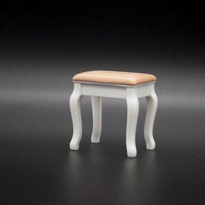 May include: A small, white vanity stool with a rectangular, light brown cushioned seat. The stool has four curved legs, and the design is reminiscent of antique furniture. The overall aesthetic is elegant and classic.
