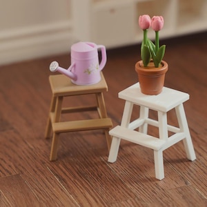 May include: Two miniature wooden step stools, one brown and one white, are displayed on a wood surface. A pink watering can with a floral design sits on the brown stool. A terracotta pot with pink tulips rests on the white stool.