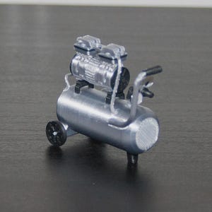 Op de afbeelding: Miniatuur zilveren luchtcompressor met een cilindrische tank, twee kleine wielen en een handvat. De compressor heeft een gedetailleerde motorcomponent aan de bovenkant en een met gaas bedekte uitlaat. Model tegen een donkere achtergrond.
