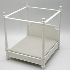 May include: A miniature white four-poster bed with a light gray mattress. The bed frame has decorative turned posts and a rectangular canopy. The headboard is solid with a curved top. The bed is designed for dolls or small figurines.