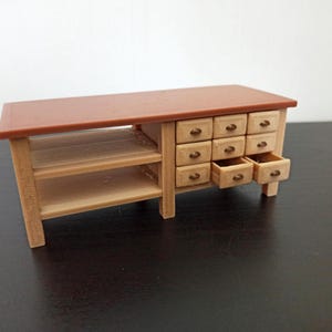 Puede incluir: Un banco de trabajo de madera en miniatura con una parte superior marrón rojiza y un marco marrón claro. Cuenta con dos estantes y una hilera de nueve cajones pequeños, tres de los cuales están abiertos. El banco de trabajo está diseñado para una casa de muñecas o un entorno en miniatura.