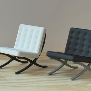 May include: Two miniature chairs, one white and one black, are displayed. The white chair has a black frame, while the black chair has a silver frame. Both chairs feature a tufted seat and a distinctive X-shaped base.