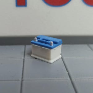 Può includere: Una batteria per auto in miniatura, con un corpo bianco e un coperchio blu. Ha due terminali argentati. La batteria è posizionata su una superficie piastrellata grigia. Lo sfondo è un muro bianco con un design blu e rosso.