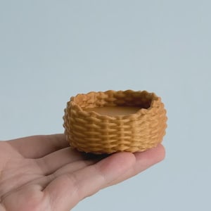 May include: A small, round, woven basket in a warm, golden-brown hue. The basket is held in a hand, showcasing its intricate weave pattern. The basket's interior is a lighter shade of brown.