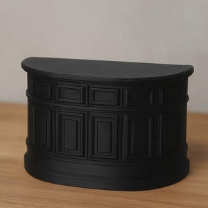 Op de afbeelding: Een zwarte, halfronde decoratieve kast met een platte bovenkant en een gedetailleerde, gepaneelde voorkant. De kast is gemaakt van een solide materiaal en staat op een houten oppervlak.