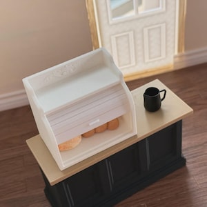 Puede incluir: Una panera blanca en miniatura con diseño enrollable, que contiene varios panes pequeños y redondos. Se encuentra sobre una encimera negra y beige, con una pequeña jarra negra cerca. La escena evoca un ambiente de cocina acogedor.