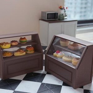 Op de afbeelding: Miniatuur bruine bakkerijvitrinekasten gevuld met diverse miniatuurgebakjes. De kasten hebben glazen fronten, met gedetailleerde miniatuur taarten, donuts en andere bakkerijproducten. De scène speelt zich af op een zwart-wit geblokte vloer.