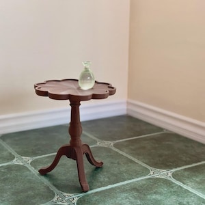 May include: A miniature brown wooden side table with a scalloped top and three legs. A small, clear glass bottle sits on the table. The table is on a green tiled floor.