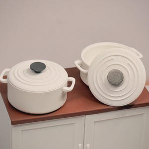 Könnte beinhalten: Weißes Keramik-Kochgeschirr-Set mit zwei Töpfen und Deckeln. Die Töpfe haben Griffe und dunkelgraue Knöpfe. Die Deckel haben ein Design aus konzentrischen Kreisen. Das Kochgeschirr wird auf einer braunen Oberfläche präsentiert.