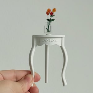Op de afbeelding: Miniatuur witte tafel met gebogen poten, met een kleine glazen vaas met oranje en rode bloemen. De tafel heeft een decoratief bloemenontwerp aan de voorkant. Een hand houdt de tafel vast.