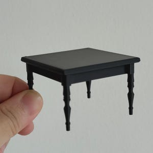 May include: A small, black, square table with four ornate legs. The table is held in a hand against a plain white background. The table's dimensions appear to be approximately 4 x 4 inches.