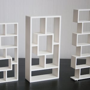 May include: Three white decorative shelves with geometric designs. The shelves are rectangular and feature open compartments of various sizes. The shelves are arranged on a dark surface against a white wall.