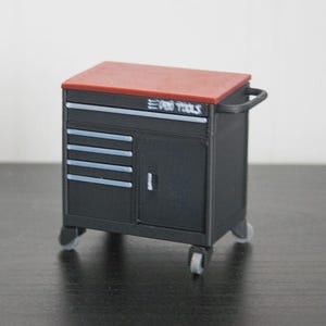 Puede incluir: Un cofre de herramientas negro en miniatura con una tapa roja y el texto "PRO TOOLS" en la parte delantera. Tiene un asa, cajones, una puerta de armario y se apoya sobre pequeñas ruedas. Ideal para un taller o garaje.