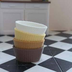 May include: A stack of four woven baskets in varying colors: white, yellow, tan, and brown. The baskets are oval-shaped and have a textured, woven design. They are stacked on a black and white checkered floor.