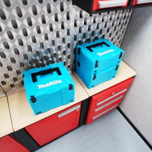 Op de afbeelding: Twee lichtblauwe Makita gereedschapskisten met zwarte handgrepen en het Makita-logo in het wit. De gereedschapskisten zijn gestapeld op een lichtgekleurd oppervlak, boven rode kasten met zilveren handgrepen. Op de achtergrond een geperforeerde metalen wand.