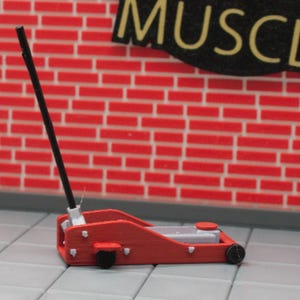 May include: A miniature red car jack with a black handle and wheels. The jack is set against a red brick wall with a sign that says "MUSCLE". The jack is a model or toy.