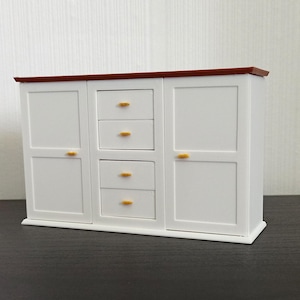 May include: A miniature white cabinet with a dark brown top and gold-colored drawer pulls. The cabinet has two doors and three drawers. The piece is designed to look like a small dresser or storage unit.