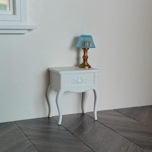 Op de afbeelding: Een witte miniatuur nachtkastje met sierlijke details en gebogen poten. Bovenop staat een kleine lamp met een gouden voet en een blauwe lampenkap. De lamp en het nachtkastje staan tegen een witte muur.