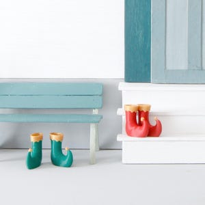 Könnte beinhalten: Ein Miniaturhaus in Türkis mit einer hellblauen Tür, weißen Stufen und einem schwarzen Briefkasten mit der Aufschrift "POST". Vor dem Haus befinden sich eine türkisfarbene Bank, ein Besen und bunte Stiefel. Das Dach ist mit kleinen bunten Fähnchen verziert.