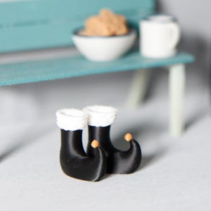 Könnte beinhalten: Miniaturszene mit einer türkisfarbenen Bank, einer Schüssel mit braunen Objekten und einer weißen Tasse. Ein schwarzer Ofen und ein dekorativer Gegenstand befinden sich im Hintergrund. Zwei schwarze Stiefel mit weißer Verzierung im Vordergrund.