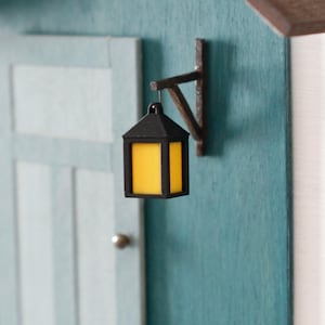 Könnte beinhalten: Miniatur-Holzhausmodell mit türkisfarbener Tür und Wand, weißen Stufen und einer kleinen Bank. Eine schwarze Laterne mit gelbem Schein hängt an der Wand. Ein grüner Stiefel mit orangefarbenem Bund steht in der Nähe der Stufen.
