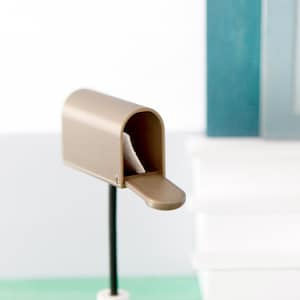 Könnte beinhalten: Eine Miniatur-Szene mit einem beigefarbenen Briefkasten mit einem Brief, einem Paar Hausschuhen und einem kleinen Besen. Der Hintergrund umfasst eine weiße Treppe und eine türkisfarbene Tür, was eine gemütliche Ästhetik schafft.