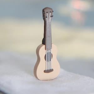 Könnte beinhalten: Eine Miniatur-Akustikgitarrenfigur in hellem Beige mit dunkelbraunem Hals und Kopfplatte, begleitet von einer kleinen schwarzen Sonnenbrille und einem Paar blau-gelber Flip-Flops, die alle auf einer weißen Stoffoberfläche liegen.