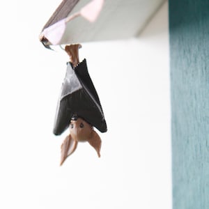 Könnte beinhalten: Eine kleine braun-schwarze Fledermausfigur, die kopfüber an der Kante einer blaugrünen Holzstruktur hängt. Die Fledermaus hat große spitze Ohren und gefaltete Flügel. Eine Schnur mit kleinen, blassen dreieckigen Wimpeln ist am Dachrand befestigt.
